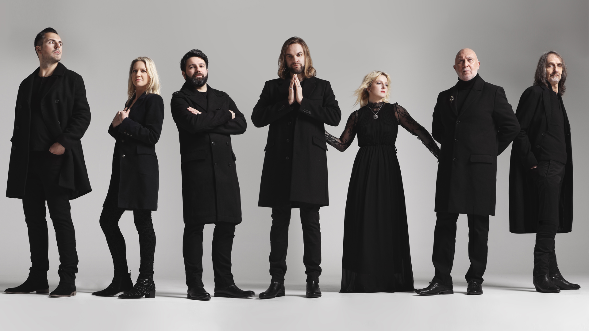 De band Rumours of Fleetwood Mac poseert voor een lichte achtergrond. Zeven leden in zwarte kleding staan naast elkaar, met variërende houdingen die een stijlvolle en dramatische uitstraling geven.